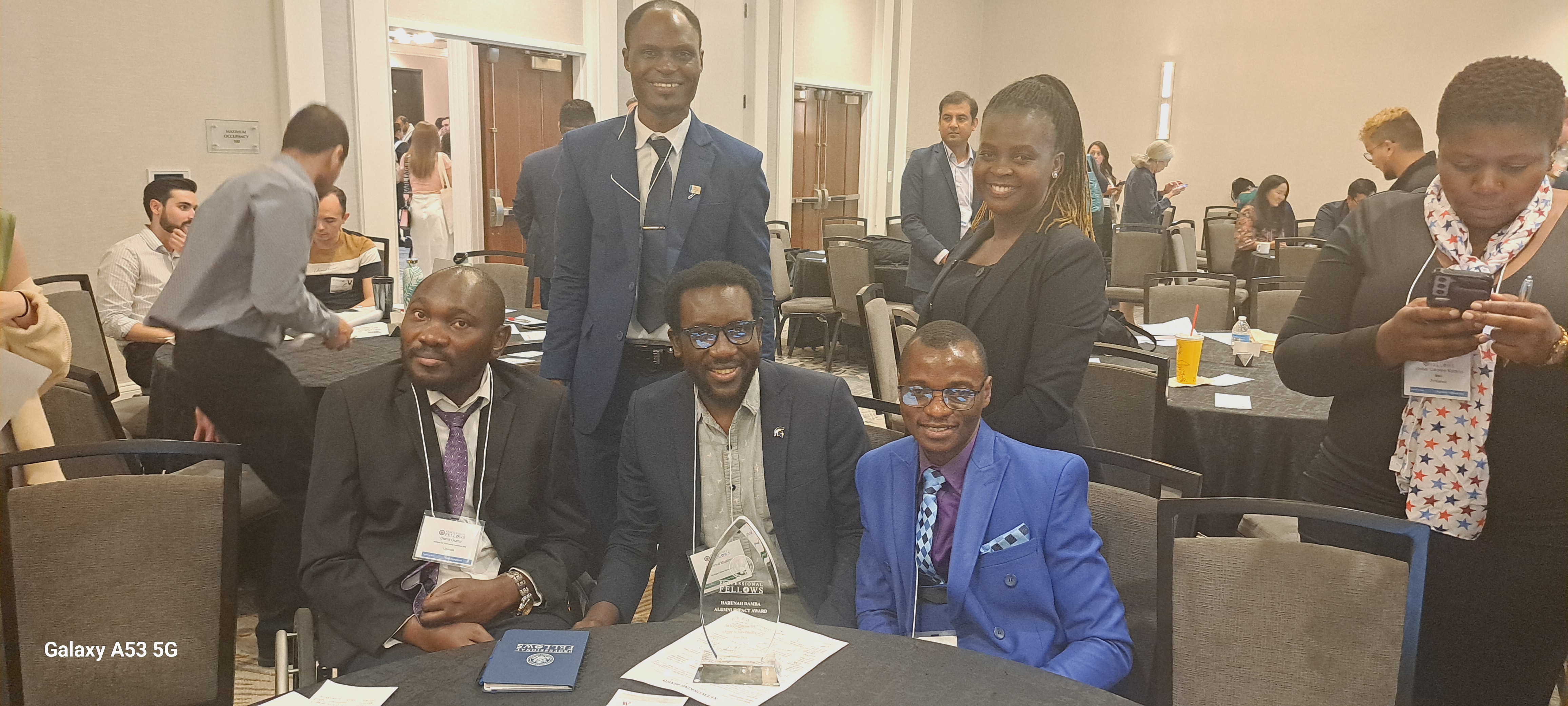 Harunah poses with colleagues during the 2024 Professional Fellows Program Alumni Impact Award ceremony in Washington, DC. His award plaque rests on the bench beside the group. From left are Denis, Raymond, and Harunah, with Moses and Stella standing behind them.